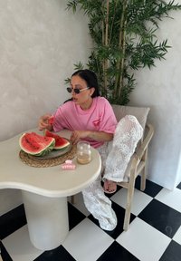 Ροζ υπερμεγέθη μπλούζα, λευκά πλεκτά παντελόνια, στρογγυλά γυαλιά ηλίου. Τραπέζι με φέτες καρπούζι, ένα μικρό βάζο και καραμέλες σε ένα υφαντό τραπεζομάντιλο.