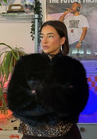 Femme aux cheveux courts bruns portant une veste en fourrure noire et un haut imprimé léopard, debout les bras croisés dans une pièce avec des plantes et des affiches de musique.