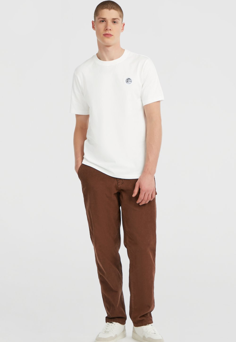 Young man wearing a white short-sleeve T-shirt with a small logo, brown trousers, and white trainers, standing with one hand in his pocket.