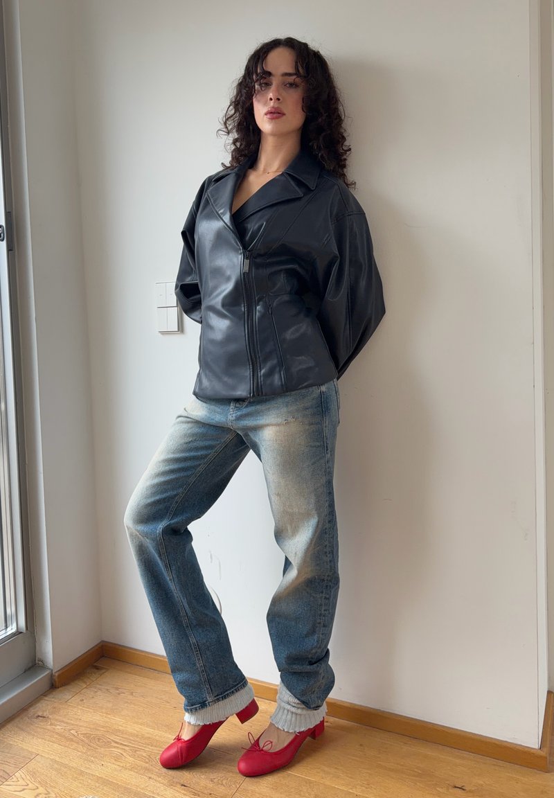 Femme aux cheveux bouclés portant une veste en cuir noire, un jean bleu délavé et des ballerines rouges, debout à l'intérieur contre un mur blanc.