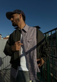 Chaqueta bomber verde sobre una camisa de cuadros, camiseta blanca y jeans de mezclilla claros. Cielo azul brillante de fondo.