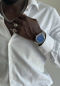 Silberne Uhr mit blauem Zifferblatt, Armband aus rostfreiem Stahl. Weißes Hemd mit Knopfdetails. Schwarze Kettenhalskette und metallische Ringe an der Hand.
