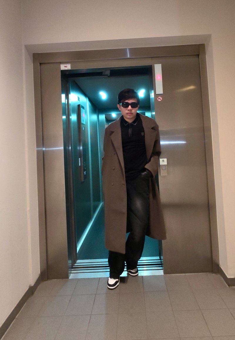 Lang brun frakke, sort polo, mørke jeans og sorte solbriller. Står ved en elevatorindgang med metalklædte vægge og blåt lys.