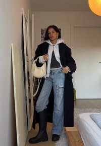 Manteau noir oversize, sweat à capuche zippé gris, haut foncé, jean bleu déchiré, bottes noires montantes jusqu'aux genoux et petit sac à main blanc avec franges.