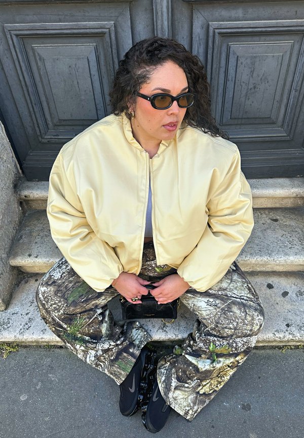 Person with curly hair wearing black sunglasses, oversized pale yellow jacket, camouflage pants, and black sneakers sitting on stone steps holding a small bag.