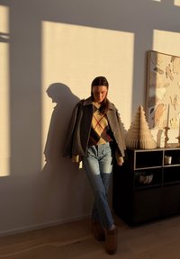 Femme debout au soleil, projetant une ombre sur le mur, portant une veste à carreaux, un pull à losanges, un jean et des bottes près d'une étagère décorative avec des œuvres d'art et des bougies.