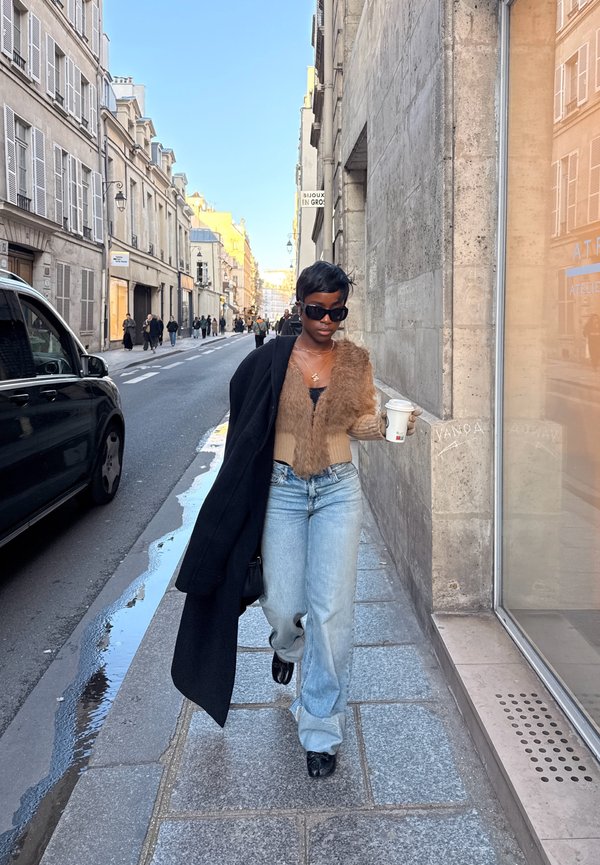 Femme portant des lunettes de soleil, un gilet en fourrure et un jean, marchant sur un trottoir en ville avec une tasse de café à la main, avec des bâtiments et des voitures le long de la rue.