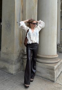 Witte gerimpelde blouse, bruine leren high-waisted flare broek en taupe plateauhakken. Model poseert tegen twee stenen zuilen.