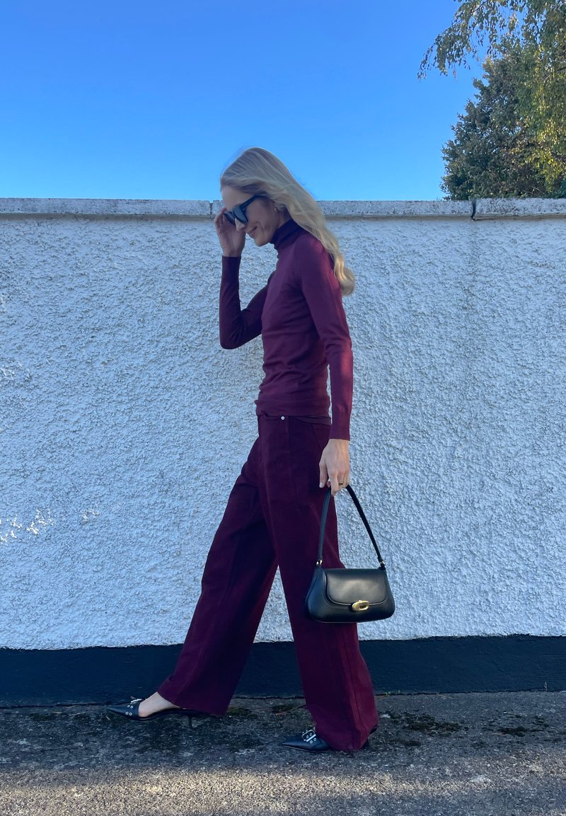 Maglione a collo alto color marrone bordeaux abbinato a pantaloni in velluto a coste larghi dello stesso colore. Borsa nera con parte superiore curva e scarpe nere a punta. Sfondo di cielo sereno.