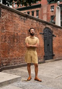 Beige polo shirt, matchande shorts och bruna sandaler; står mot en tegelvägg med en svart dörr och grönska ovanför.