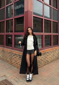 Schwarzer Trenchcoat über einem weißen Hemd, grauen Shorts und schwarzen Mary Jane Schuhen. Die Accessoires umfassen Sonnenbrillen und eine kleine schwarze Handtasche.
