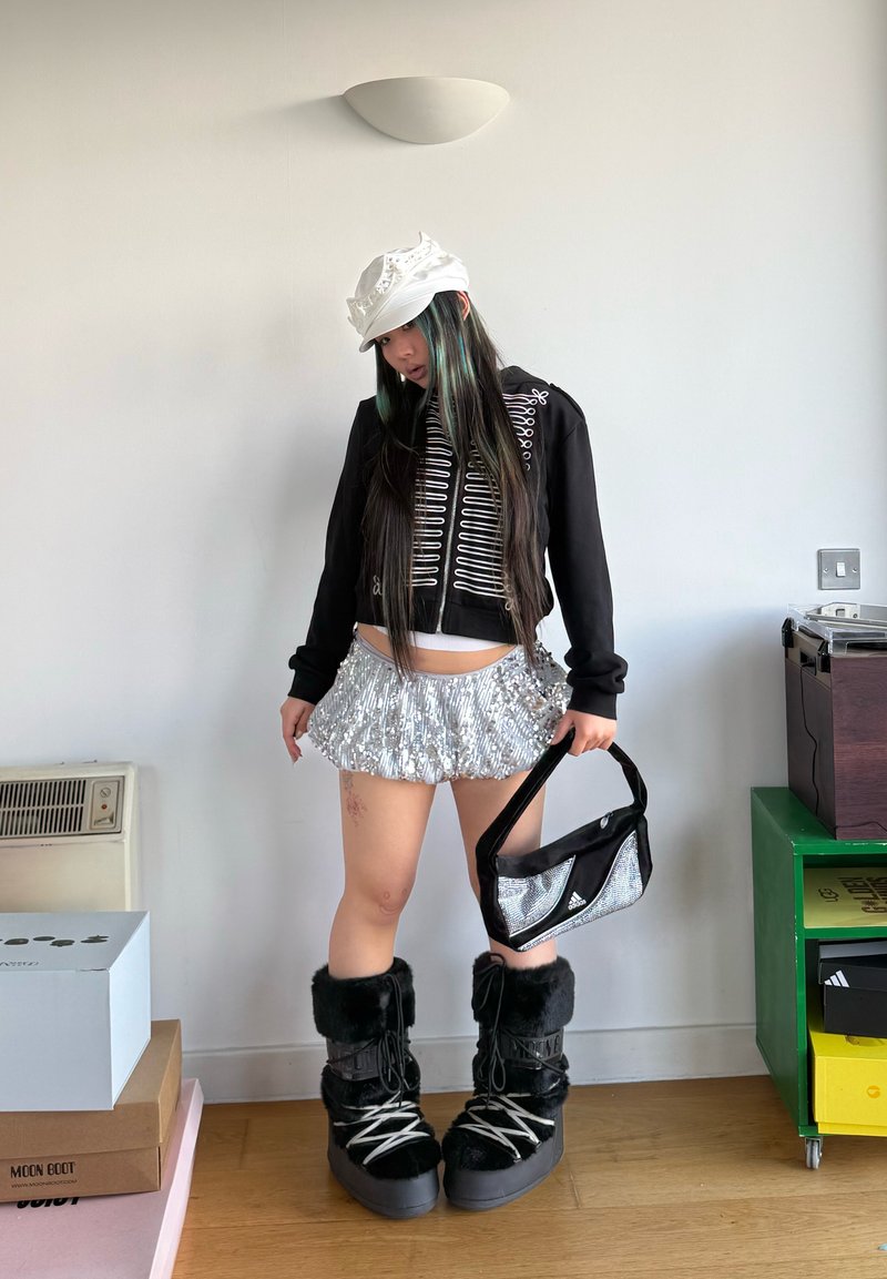 Jeune femme portant une casquette blanche, une veste noire avec une bordure blanche, une jupe en sequins argentés, tenant un sac Adidas noir et argenté, et des bottes en fourrure noires à l'intérieur.