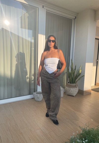 Gafas de sol negras, top strapless blanco y pantalones sueltos beige de estampado. De pie sobre un suelo de baldosas con plantas en macetas y reflejos de luz solar.