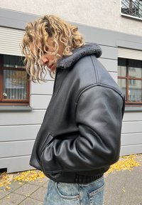 Must nahast jakk halli karvakaelusega, mis on tekstuurse pinnaga, üle suurusega ja soonikkoes äärega, kombineeritud helesiniste teksadega.