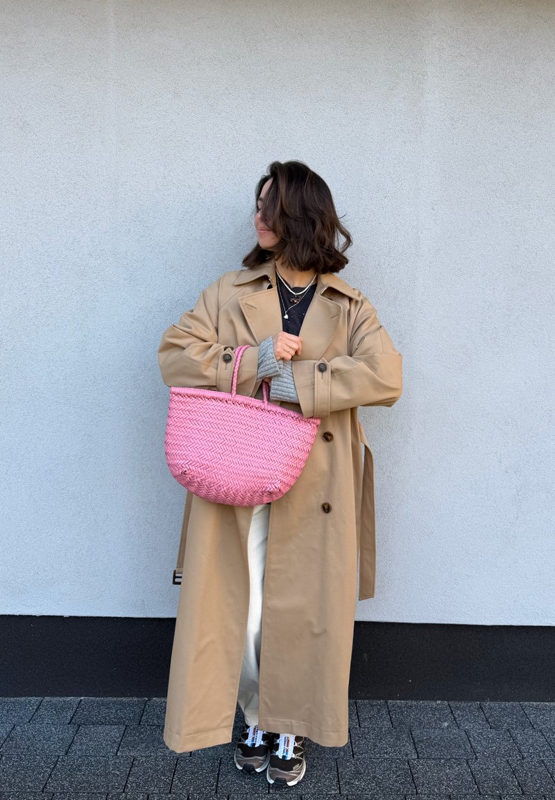 Kvinna som bär en lång beige trenchcoat och vita byxor, håller en stor rosa flätad väska, stående mot en enkel grå vägg.