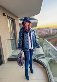Chaqueta de cuero negra, leggings azul marino, botines teal, bolso gris, sombrero negro, gafas de sol extragrandes; de pie en un balcón con vistas al atardecer.