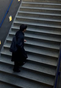 Szürke kőlépcsők jellegzetes textúrákkal, kék korláttal körülvéve. Egy fekete kabátot viselő személy felmegy, sötét hátizsákot cipel.