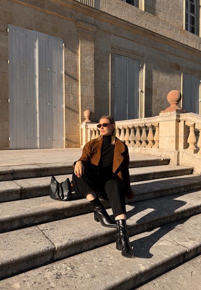 Chaqueta de ante marrón, cuello alto negro, pantalones negros, botas de tacón y un bolso negro. Sentada en unos escalones de piedra con luz solar y sombras.