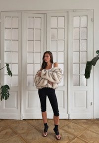 Fuskepelsjakke i beige og krem, med en fluffy tekstur. Svart, tettsittende leggings og svarte sandaler med stropper. Glassdører i bakgrunnen.