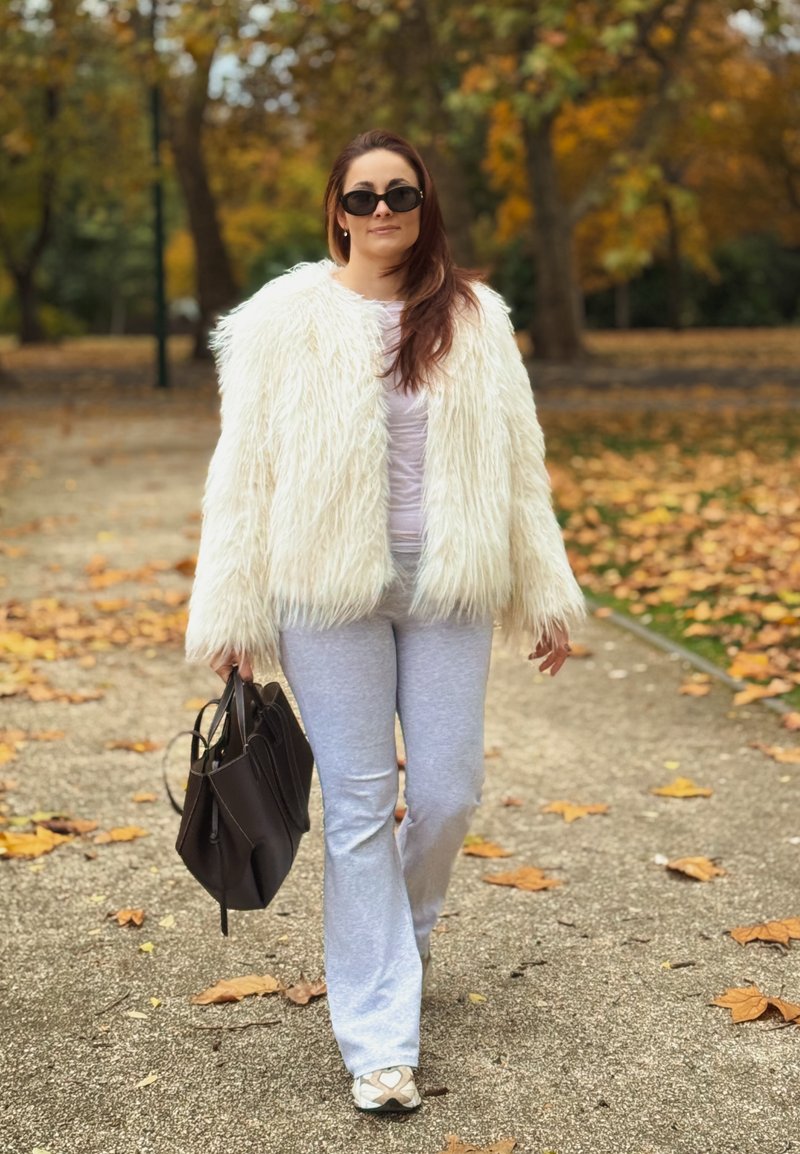 Bijela lažna krznena jakna, svijetla majica, sivi hlače, crna torba. Naočale za sunce na licu. Ležeran, teksturiran outfit među padajućim jesenskim lišćem.