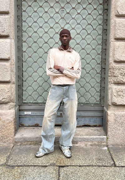 Chaqueta rosa claro con cremallera y acentos marrones, rayas blancas; jeans de mezclilla azul desgastados y oversize; zapatillas deportivas plateadas; fondo de malla gris.