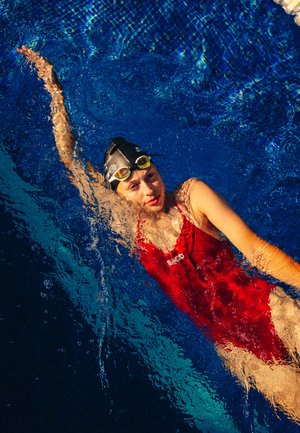 Traje de baño rojo con un escote alto, acompañado de gafas de natación negras. El sujeto está parcialmente sumergido en agua azul clara.