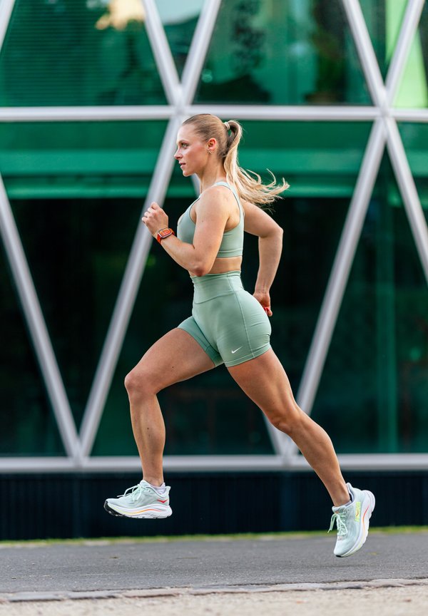 Vrouw in groene sportkleding loopt buiten voorbij een gebouw met geometrische raam patronen, haar in een paardenstaart, geconcentreerde uitdrukking.
