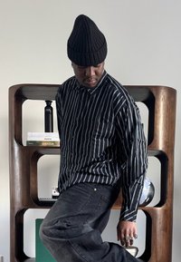 Homme portant un bonnet noir et une chemise rayée, posant devant une étagère en bois avec des livres, un globe et une bouteille d'eau.