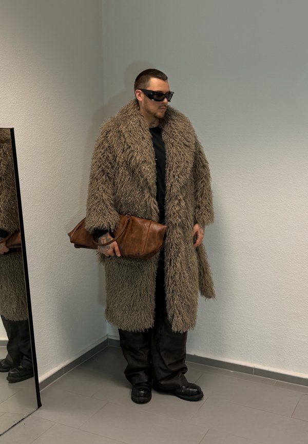 Un homme portant des lunettes de soleil, un manteau brun hirsute, des vêtements noirs et des chaussures épaisses tient un sac en cuir brun près d’un miroir.
