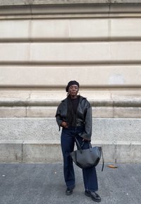 Zwarte leren jas, zwart shirt met lange mouwen, donkerblauwe jeans met wijde pijpen, zwarte tas met gebogen vorm en zwarte plateauzolen tegen een lichte muur.