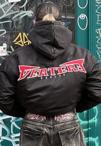 Μαύρο bomber μπουφάν με κεντημένο το κείμενο "VERTERI BERLIN" σε κόκκινο και λευκό στην πλάτη. Γκρι φούτερ από κάτω, ορατά τζιν.
