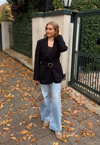 Sacou negru cu o curea, blugi largi de culoare albastru deschis și adidași bej. Ține în mână o geantă bej texturată. Frunze de toamnă împrăștiate pe trotuar.