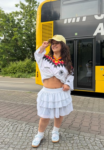 Joven mujer con falda blanca de capas y camiseta deportiva ajusta una gorra amarilla mientras está de pie en la acera cerca de un autobús amarillo.
