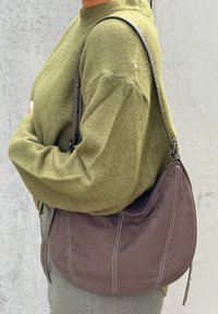 Barna velúrbőr válltáska, kontrasztos fehér varrással, ívelt formával és állítható pánttal, olíva zöld pulóver mellett viselve.