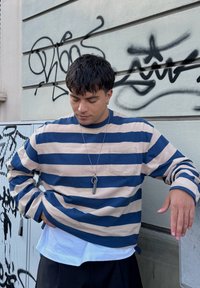 csíkos, bézs és sötétkék hosszúujjú ing, mellzsebkával. Fehér ing fölé rétegezve, sötét nadrággal párosítva, nyaklánc medállal.