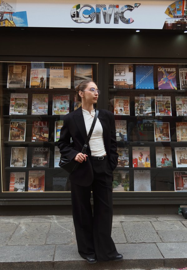 Donna in completo nero e occhiali in piedi su un marciapiede davanti a una vetrina di riviste etichettata "CIVIC".