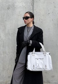 Hvid håndtaske med lynlåslukning, frontlomme og dekorativt beslag; kombineret med en sort frakke og gråt outfit. Glat tekstur.