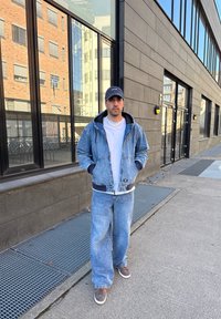 Denimjakke med snorhette, marineblå detaljer, lyseblå jeans, hvit t-skjorte og brune sko. Står utenfor en moderne bygning.