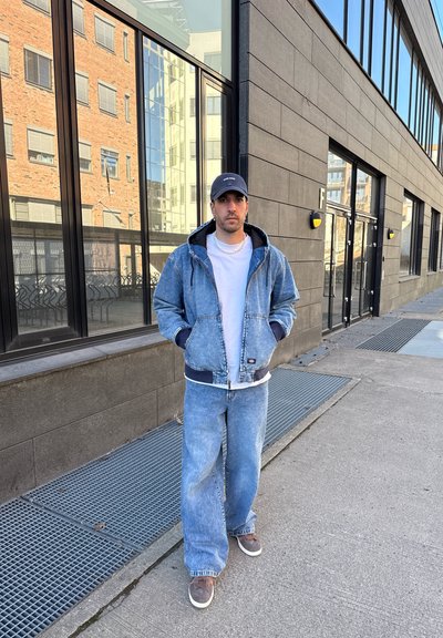 Chaqueta de mezclilla con capucha ajustable, acentos en azul marino, jeans de color azul claro, camiseta blanca y zapatos marrones. De pie frente a un edificio moderno.