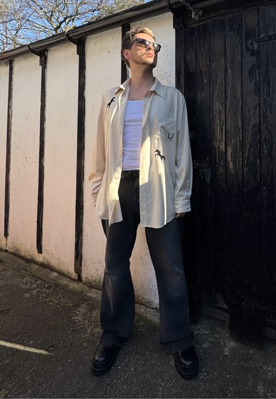 Camisa crema con bordado de caballo, camiseta sin mangas blanca, pantalones de pierna ancha negros y botas negras gruesas. De pie contra una pared y puerta texturizadas.