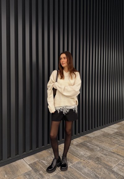 Mujer de pie junto a una pared de listones verticales negros, vistiendo un suéter crema de gran tamaño, pantalones cortos negros con dobladillo de encaje, medias negras y zapatos negros.