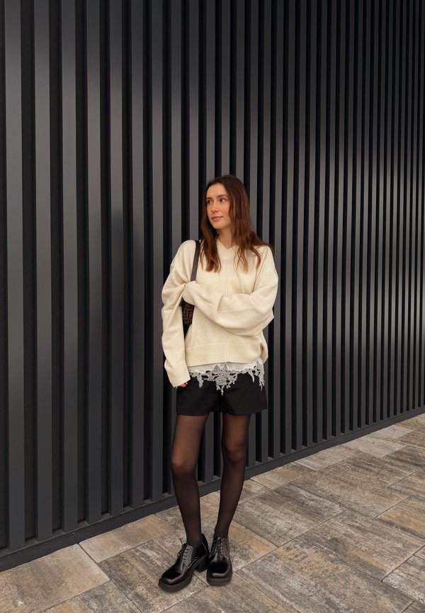 Vrouw staand bij een zwarte muur met verticale latten, gekleed in een crèmekleurige oversized trui, zwarte korte broek met kanten zoom, zwarte panty's en zwarte schoenen.