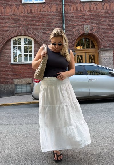 Top sin mangas negro, falda larga blanca con volantes, bolso de paja marrón trenzado, chanclas negras. Fondo de edificio de ladrillo con ventanas en arco.