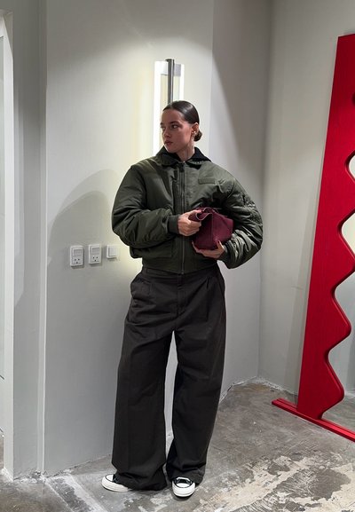 Chaqueta bomber verde, pantalones negros, sosteniendo un bolso burdeos. Zapatillas blancas, textura suave y diseño minimalista en un interior moderno.