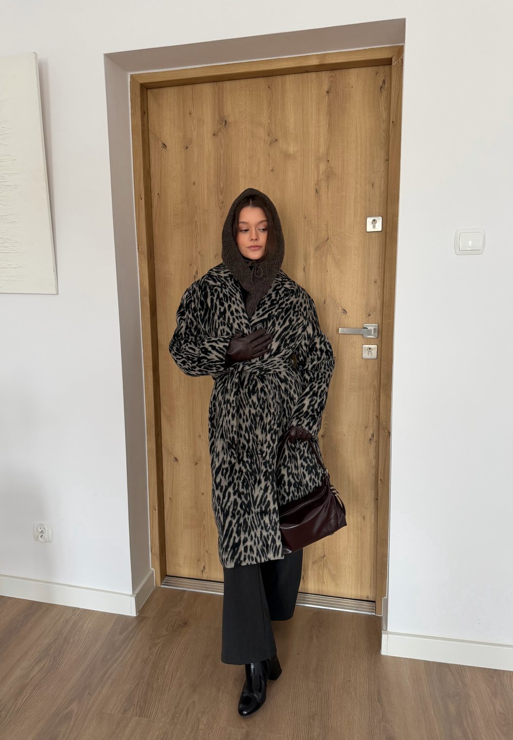 Manteau à motif léopard style fourrure avec manches larges, écharpe en laine brune, pantalon foncé et sac à main bordeaux, debout près d'une porte en bois.