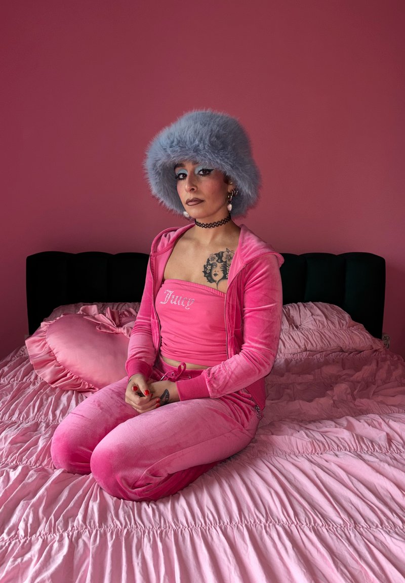 Pinkfarbener Velours-Trainingsanzug mit "Juicy"-Logo, flauschiger hellblauer Hut, schwarzes Halsband, sitzt auf rosa Rüschenbettwäsche mit herzförmigem Kissen.