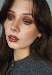 Nærbilde av en models ansikt med brune øyenskygger, en gyllen septumring, glossy rødlig lepper, og iført en grå teksturert genser.