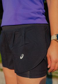Osoba u crnim slojevim sportskim kratkim hlačama i ljubičastoj majici, s pametnim satom na zapešću, stoji na otvorenom.