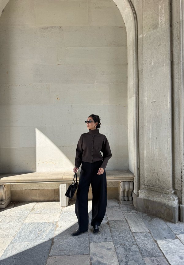 Donna con maglione scuro, pantaloni larghi neri, occhiali da sole, tiene una borsa nera, in piedi in un arco di pietra con panchina al sole e all'ombra.
