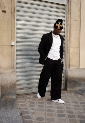 Blazer negro sobre una camisa blanca, combinado con pantalones negros holgados y zapatillas blancas. Un gorro negro texturizado presenta un diseño de cruz colorido.